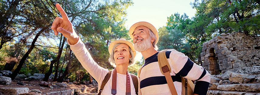 A couple in their early sixties are standing in front of ruins. There are many trees surrounding the ruins and the sun is just behind the couple. They are both wearing hats and backpacks, the woman has a camera around her neck.