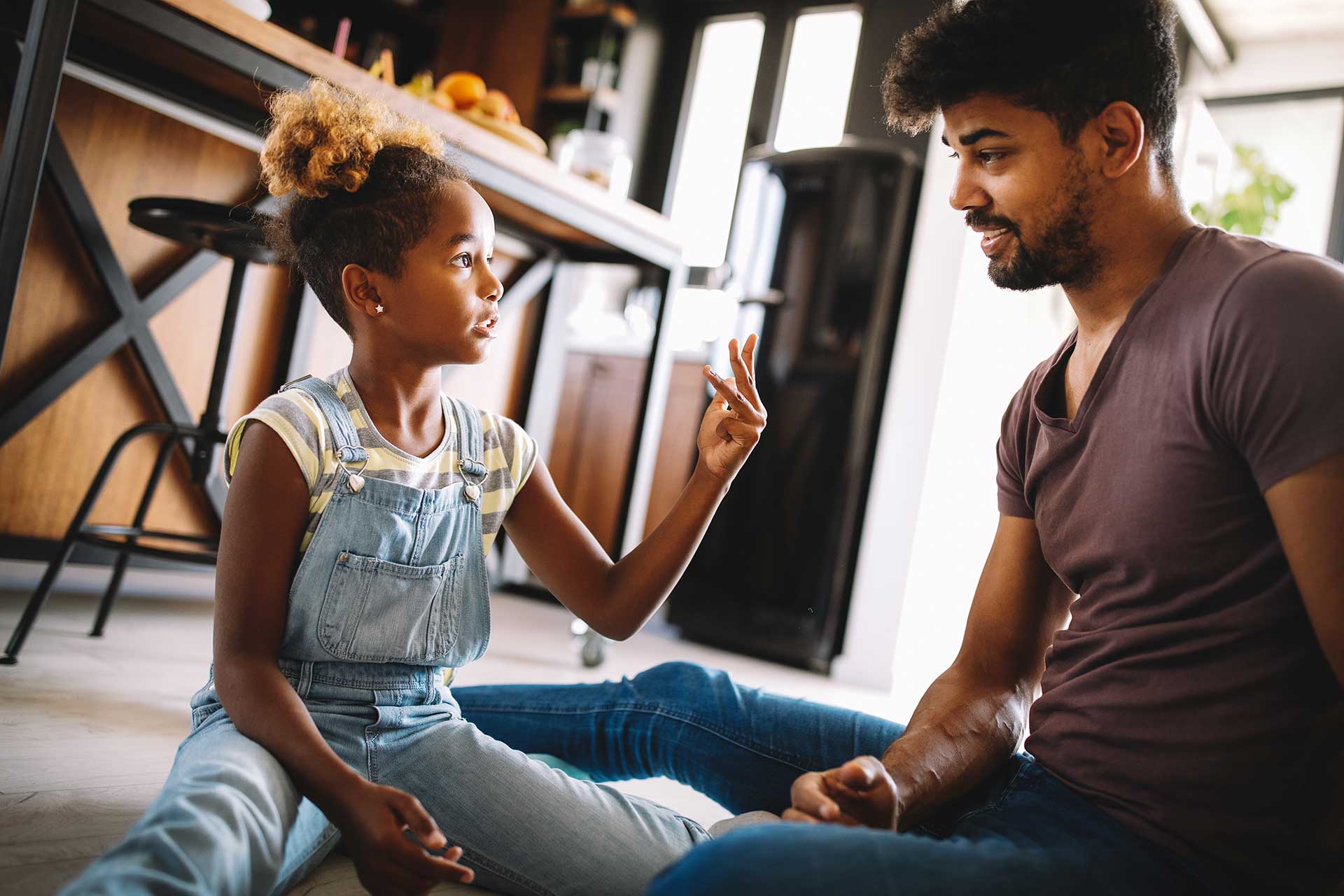 A father is talking his young daughter. The are both sitting on the floor, the daughter looks concerned and he is comforting her.